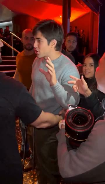 The man in the grey sweater stands in the red-lit venue among the crowd