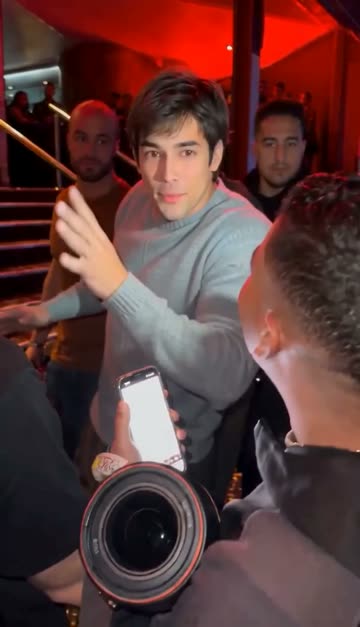 The man in the grey sweater gestures with his hand while people film him